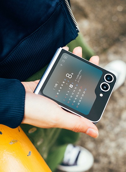折りたたみスマホ「Galaxy Z Flip6(ギャラクシーZフリップ6)」を、カレンダーが写ったカバー画面を上にして片手で持っている。