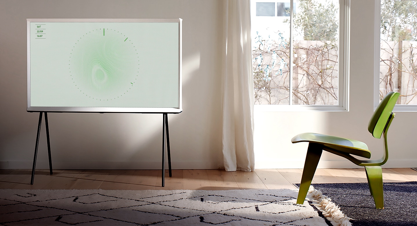 A Cloud White model of The Serif sits on a black stand in a bright and elegantly simple living room, with an area rug, a stylish three-legged green chair, and large windows. The image on screen is Leaf Ambient Mode with clock ticking.