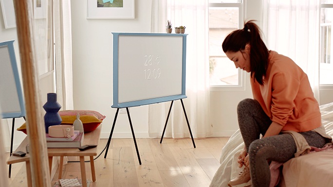 A woman sits on her bed tying her shoelaces. A Cotton Blue model of The Serif rests neatly on a tv stand in the corner with time and weather information on the screen using Ambient mode.