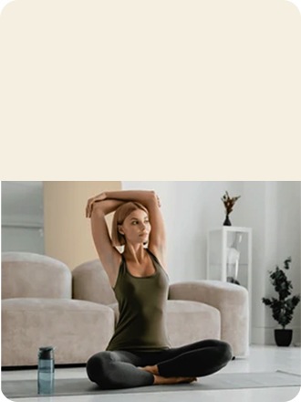 Una mujer se estira sobre una colchoneta de yoga con los brazos levantados en la sala de estar. Detrás de ella, un sofá beige y una planta en maceta.