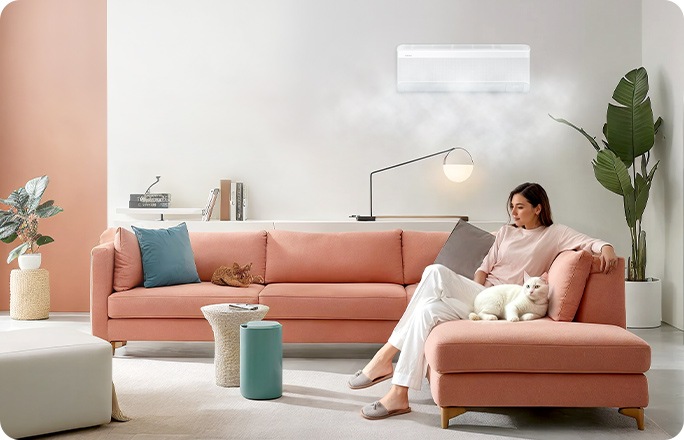 Un suave chorro de aire sale de un aparato de aire acondicionado Samsung windfree instalado en la pared del salón. Una mujer está cómodamente sentada en el sofá con un gato.