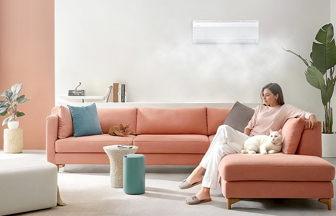Un suave flujo de aire sale de un aire acondicionado Samsung WindFree montado en la pared de una sala. Una mujer está cómoda sentada en un sofá con un gato.