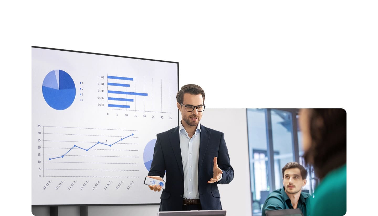 A man is giving a presentation in front of people in an office. Behind him, a large display is showing various graphs and he is holding his Galaxy smartphone in his hand.