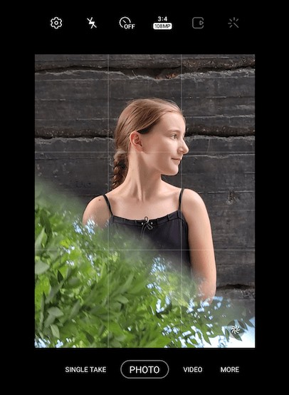 The screen of a Galaxy S21 Ultra 5G  showing a girl wearing a black shirt looking off to her side with green leaves reflected on the bottom half of the photo