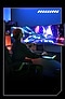 A woman is seen from the back, fully immersed in a game. Her right hand is placed on a mouse, which is on a mousepad that radiates blue-green lights. A horizontally placed Odyssey Ark Monitor is seen from the front and shows a spaceship dashing through a gateway. Blue-green beams are seen on the edges of the spaceship, indicating its interstellar speed. A moon and a few urban buildings are partially seen in the background.