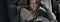 A woman in a beige sweater, sitting on her couch while holding her Galaxy phone. Her body is turned toward the wall and she is looking out the window as her left hand supports her head.