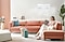 A gentle airflow comes from a Samsung windfree air conditioner mounted on a living room wall. A woman sits comfortably on a sofa with a cat.