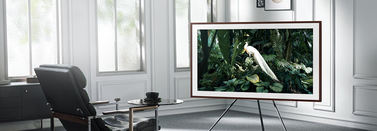 A Samsung lifestyle TV sits on a three-legged stand in front of a black leather armchair in a white room with large windows. There is an image of a large white bird on the screen.