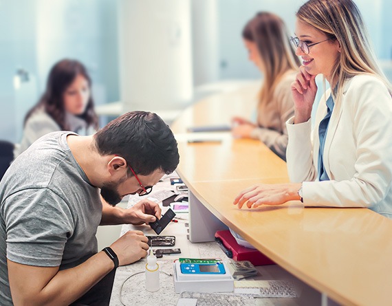 A man is scrutinizing the disassembled parts of a phone, with various tools spread out on the long counter before him. Across the counter, a smiling woman observes the process. In the background, another woman is being serviced by another tech worker.
