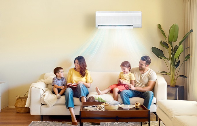 Fast, cool air flows from a Samsung windfree wall-mounted air conditioner in a living room with a family of four sitting on a beige sofa. The parents and two children are enjoying their time together.