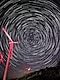 Long exposure shot of the stars in the night sky, forming a circular pattern that fill the sky. There are six red wind power generators in the foreground facing the left.