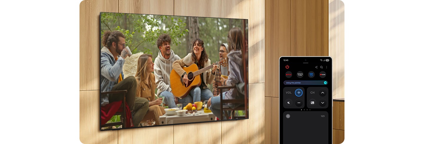 A Samsung Smart TV mounted to a wall displays a scene of musicians playing outdoors. A nearby smartphone that looks like a touchable remote control is used to turn up the TV volume. When the volume button is pressed on the smartphone remote control, the TV speaker volume is turned up in response on the TV.