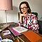 A short-haired woman with glasses sitting at her desk smiling wearing a white blouse with a red and blue pattern. On her desk are art materials and there is a pink lamp in the background.