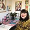 A short-haired woman with dark hair is sitting at her desk smiling wearing a leopard print top. On her desk are art materials and there are colourful art prints in the background.