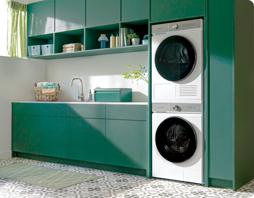 Veļas mazgājamā mašīna un žāvētājs baltā krasā ir novietoti vertikāli gaišā saimniecības telpā, kurā atrodas zaļas krāsas skapīši.