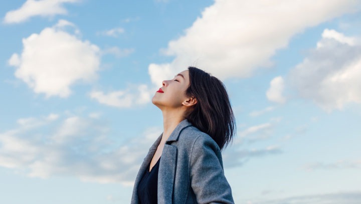 Una mujer con una chaqueta sport azul y gris está parada con la cabeza inclinada hacia un cielo azul con nubes blancas. Tiene los ojos cerrados y parece estar inhalando profundamente.