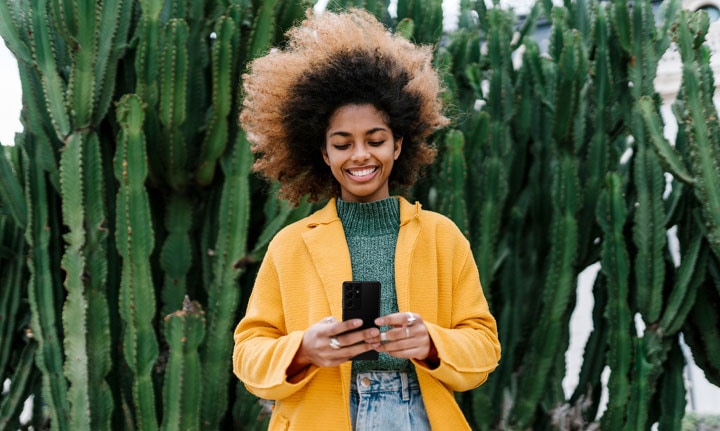 Una mujer con un abrigo brillante y el cabello al viento está parada frente a un cactus y sonríe a su Galaxy S21 Ultra.