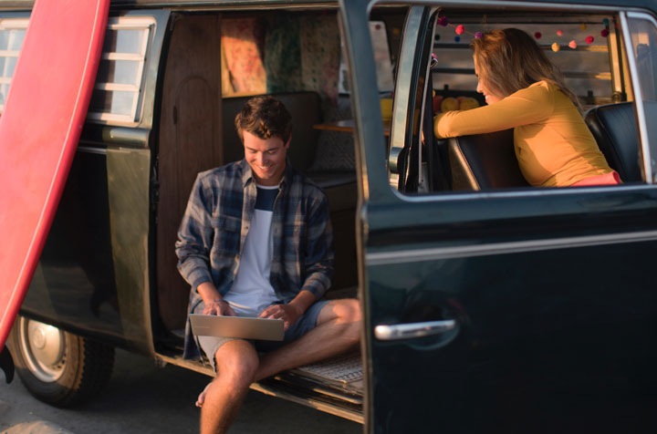 Un hombre y una mujer sentados dentro de una camioneta negra estacionada y con las puertas abiertas. El hombre sonríe mientras escribe en un Galaxy Book gris que tiene en el regazo. Sus piernas cuelgan fuera de la camioneta. Lleva una camisa de franela de manga larga y pantalones cortos. La mujer sonríe mientras gira para mirarlo desde el asiento del pasajero. Lleva una camiseta amarilla de manga larga.