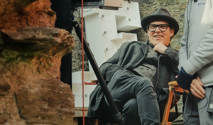 Joe Wright está sentado en una silla de director en un escenario de producción; mira un monitor y sonriendo.