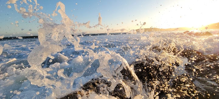 Primer plano de una ola que golpea contra las rocas en el centro de la imagen. El agua salpica frente a la cámara, iluminada por el sol que cuelga bajo en el cielo, justo fuera de cámara.