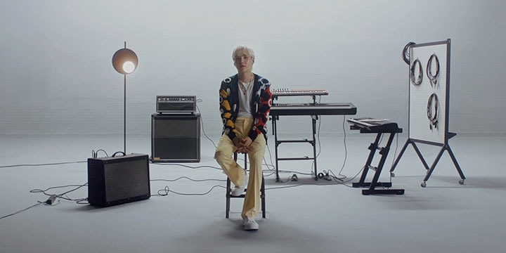 SUGA of BTS sitting in a stool, standing in the middle of an empty music studio. He’s surrounded by musical instruments. There is a speaker on the left side of the room and three electronic keyboards on the right.
