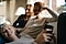 Two women are smiling as they sit on a couch. The one on the left has her left arm bent, supporting the side of her head. They are both looking down at a younger woman lying horizontally across their laps while holding up a black smartphone.