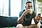Man is sitting on a couch in front of a large window. He is holding a mug close to his face as if he is about to sip a beverage while checking his smartphone with the other hand.
