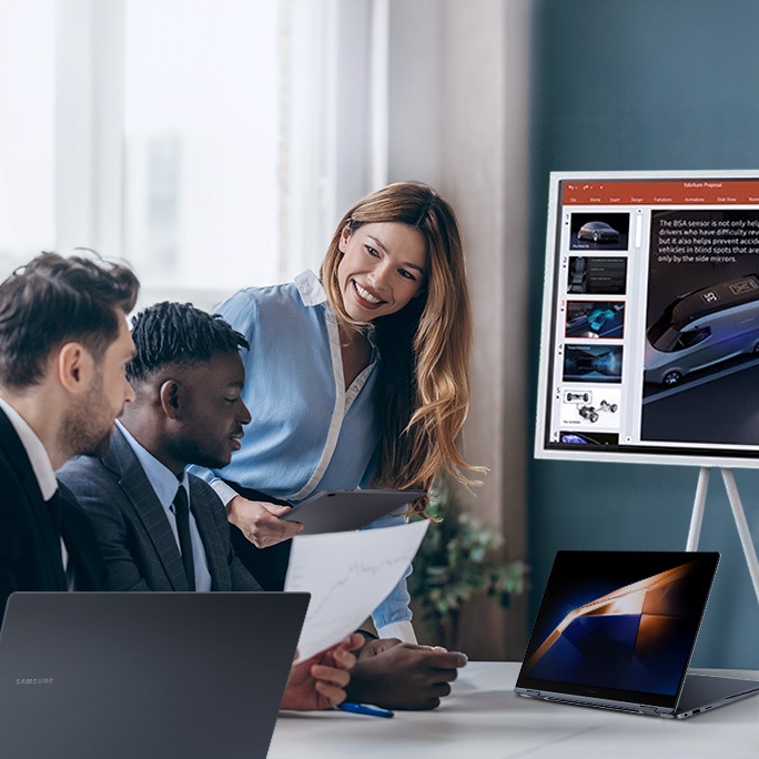 Three professionals are gathered around a table in an office setting, engaged in a team discussion. One person is holding a document, another is using a laptop, and a third is holding a tablet while standing and smiling at her colleagues. In the background, a large monitor displays a presentation slide featuring images and text. The text on the image reads "Essentials for teams." The atmosphere appears collaborative and productive.