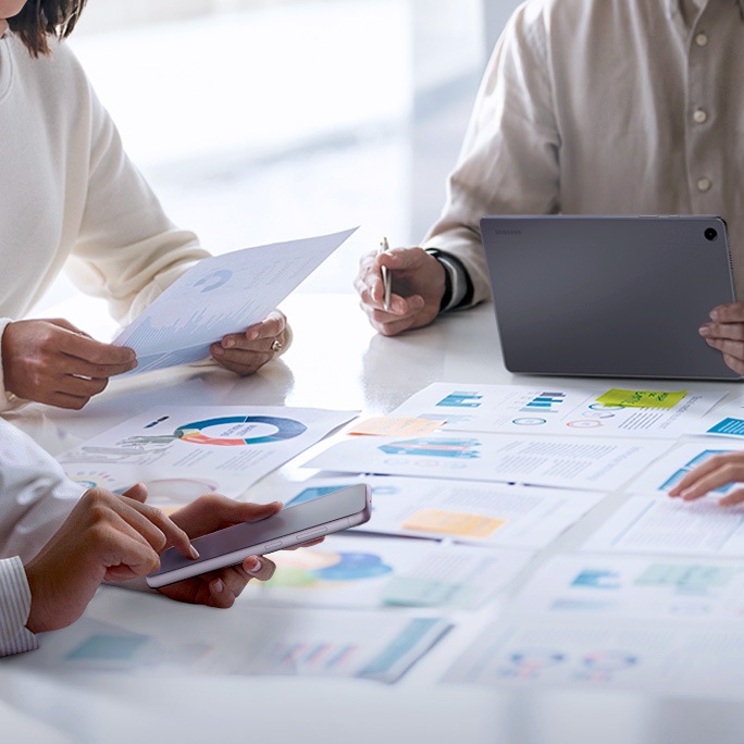 Three individuals are sitting at a table, engaged in a collaborative discussion. They are examining various charts, graphs, and documents spread out on the table. One person is holding a tablet, another is holding a document, and the third person is using a smartphone. The text on the image reads "Smart deals, bigger savings." The setting appears to be a bright, modern office environment.
