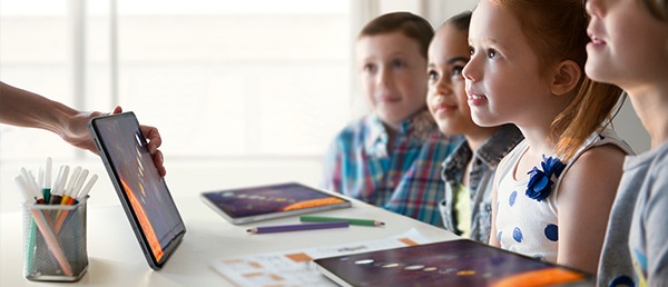 Four children with two Galaxy Tabs placed on the table, listen to and look at the Galaxy Tab screen shown by their teacher.