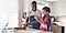 A father wearing a blue apron is baking next to his son who is wearing a red apron. His daughter is on the right side of the father clapping while father and son are making dough on a granite kitchen countertop.