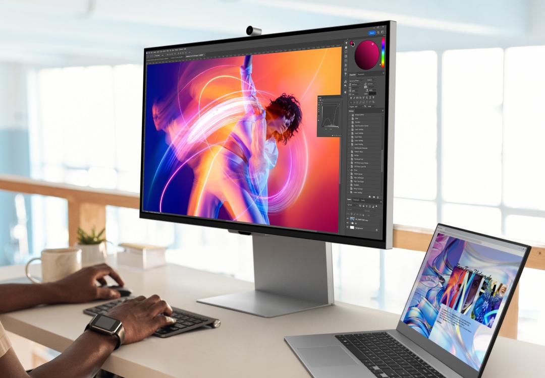 A person sits at a desk with a Samsung ViewFinity monitor displaying a vibrant image of a dancer in an editing interface. The monitor is mounted on an Easy Setup Stand, surrounded by a keyboard, mouse, and a smaller laptop on the right.