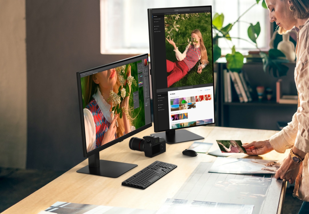 A person stands at a desk with dual Samsung ViewFinity monitors, one in landscape and the other in portrait mode, displaying vibrant images in a photo editing interface. A camera, keyboard, and mouse are on the desk, showcasing a productive, ergonomic setup.