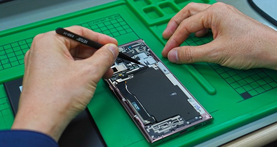 Closeup of the hands of a technician repairing a phone on a table. The phone has been taken apart with the interior components exposed