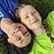 An aerial view of two women laying side-by-side on the grass. The woman on the left is wearing a blue collared shirt while the woman on the right is wearing a lime green top. Their heads are pressed together from opposite ends. They're both smiling at the camera.