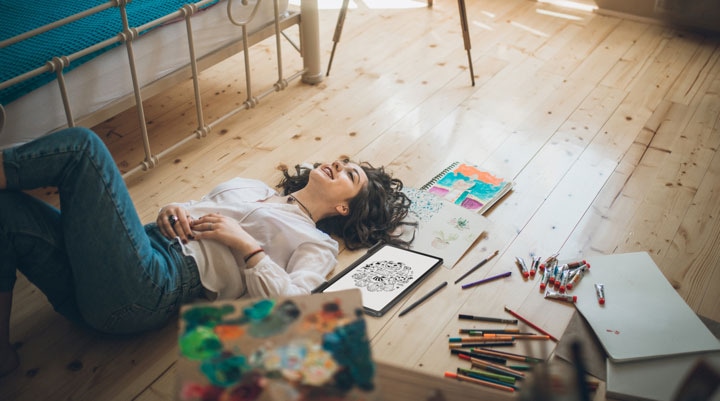 Een vrouw ligt met het gezicht naar boven op een houten vloer, markeerstiften, potloden, verf en andere kunstbenodigdheden om haar heen.