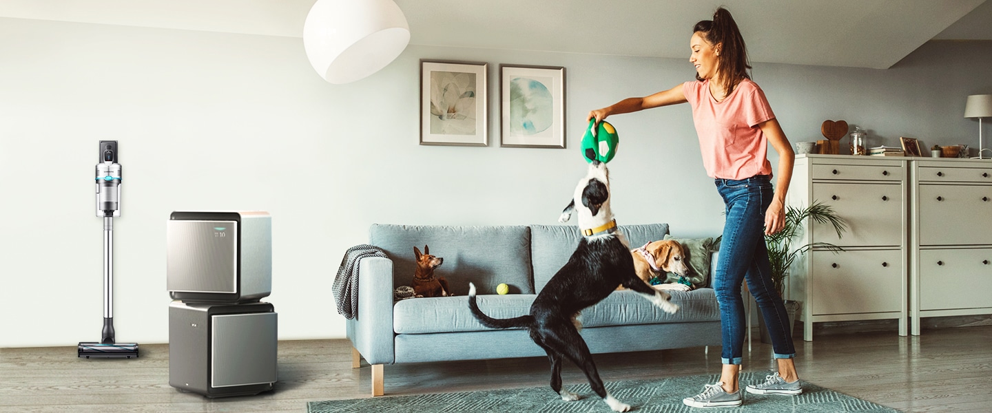 Samsung Jet 90 Pet and Samsung Cube Air Purifier are placed in a living room. Two dogs are sitting on a couch and a woman is playing catch with one other dog and a toy ball.