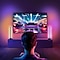 Een persoon die in een schemerige kamer zit en een energiek racevideospel speelt op een groot tv-scherm. De kamer wordt verlicht door kleurrijke sfeerverlichting, waardoor de gaming-ervaring wordt verbeterd.