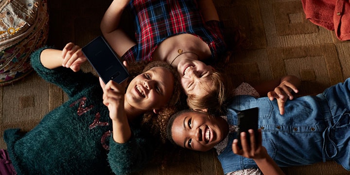 Tre unge jenter ligger på ryggen og leker med smarttelefonene sine.