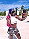 A woman with blue hair, white tanktop and jean shorts poses for a selfie.