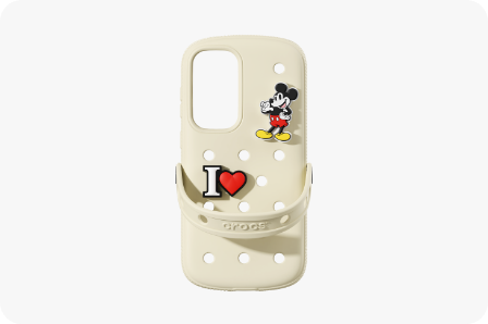 Sin el dispositivo Galaxy S25+, se coloca una Crocs Case en  beige con la parte posterior mirando hacia arriba. En la esquina superior derecha de la caja, hay una figura de Mickey Mouse, y en el lado izquierdo, la letra “I” y un corazón rojo se muestran juntos. Similar al calzado Crocs, el estuche tiene agujeros