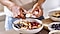 A close-up of a hand slicing a banana with a knife into a salad bowl filled with fruit in the kitchen