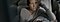 A woman in a beige sweater, sitting on her couch while holding her Galaxy phone. Her body is turned toward the wall and she is looking out the window as her left hand supports her head.