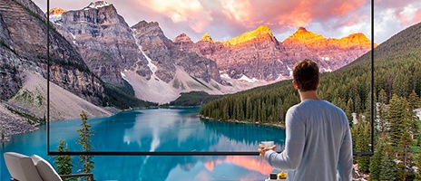 A man holding a coffee cup stands in front of a Supersize TV which shows an outdoor landscape scene of a mountain range and lake. The image expands beyond the screen.