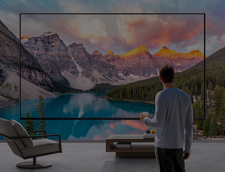 A man holding a coffee cup stands in front of a Supersize TV which shows an outdoor landscape scene of a mountain range and lake. The image expands beyond the screen.