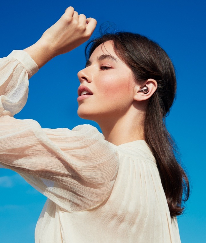 A young woman wearing a white blouse is shown from her left profile with a Galaxy Buds Live earbud in her left ear.