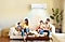 Fast, cool air flows from a Samsung windfree wall-mounted air conditioner in a living room with a family of four sitting on a beige sofa. The parents and two children are enjoying their time together.