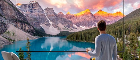 رجل يحمل فنجان قهوة يقف أمام تلفزيون ضخم جدًا يعرض منظرًا طبيعيًا لسلسلة جبال وبحيرة. تتسع الصورة خارج الشاشة.