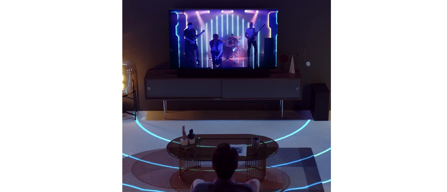 Uma televisão QLED numa sala escura transmite um concerto. Representação gráfica dos sons emitidos e optimizados para o espaço pela soundbar Q Series Samsung.