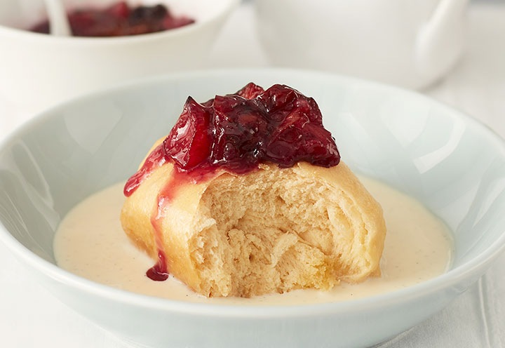 Num prato branco está um bolinho com compota de ameixa assada em cima. E o creme de baunilha está disposto à volta do bolinho.